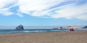 Large Rock in the ocean, blue sky and clouds beyond, sandy beach and red car in foreground.