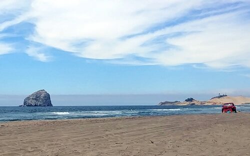 Large Rock in the ocean, blue sky and clouds beyond, sandy beach and red car in foreground.