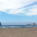 Large Rock in the ocean, blue sky and clouds beyond, sandy beach and red car in foreground.